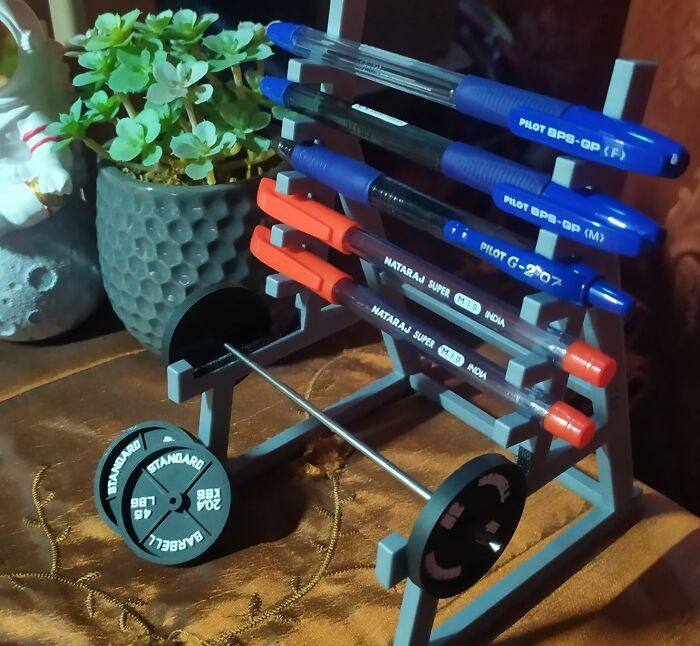 Pens displayed on a desk with a weightlifting-themed holder, next to a plant, enhancing work supplies aesthetics.