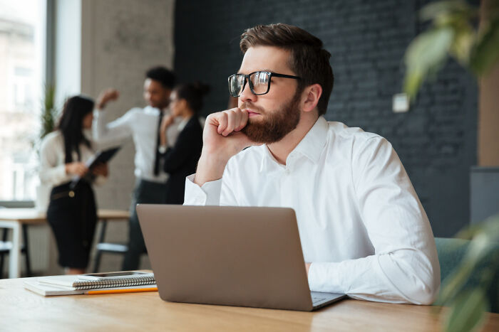 Man in glasses deep in thought at a desk, with colleagues discussing in the background, focusing on what keeps him up at night.
