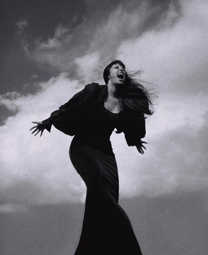 A woman in a long dress, captured in a dramatic pose against a cloudy sky, showcasing dreamy dark escapism photography.