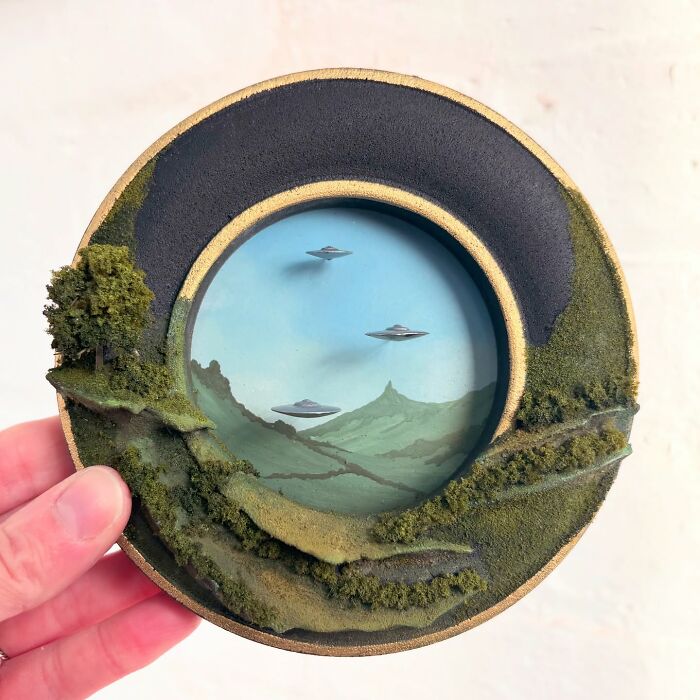 Hand holding a diorama of UFOs flying over a mysterious landscape, surrounded by mossy textures.