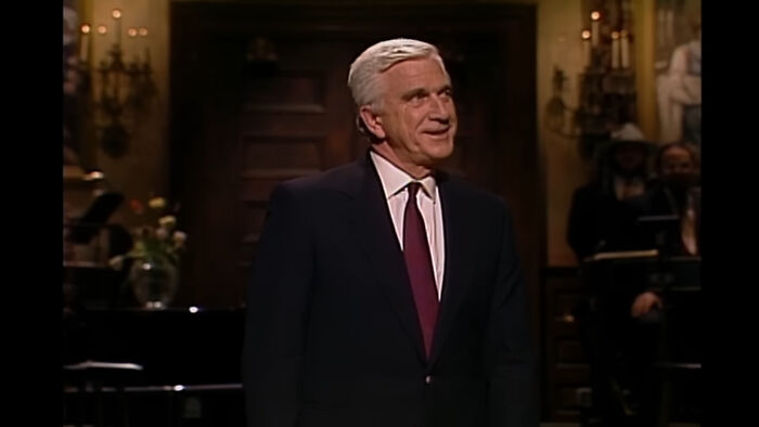 Elderly man in a suit on stage with a classic interior backdrop, embodying the charm of popular celebrities.