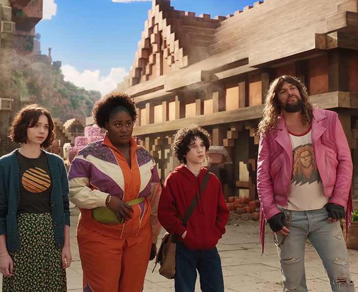 Four people standing in a 'Minecraft' scene with wooden buildings, one in a pink jacket and jeans.