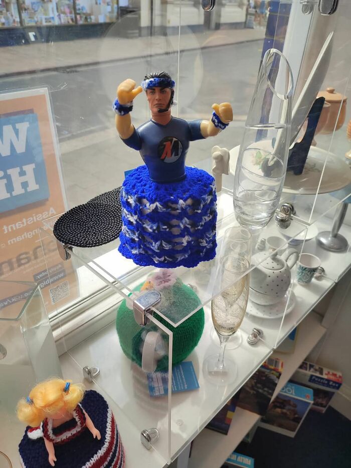 Bizarre treasure in a secondhand shop: action figure with blue crochet skirt, displayed alongside mugs and glasses.