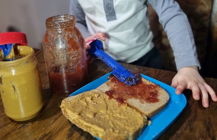 Child spreading jam on bread, next to peanut butter jar on table; a genius kitchen find for group chat sharing.
