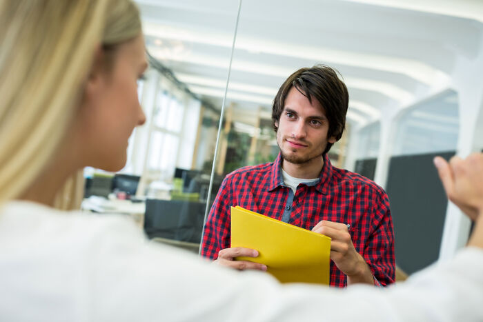 Young man in a red plaid shirt holding a folder during a job interview answering ridiculous interview questions.