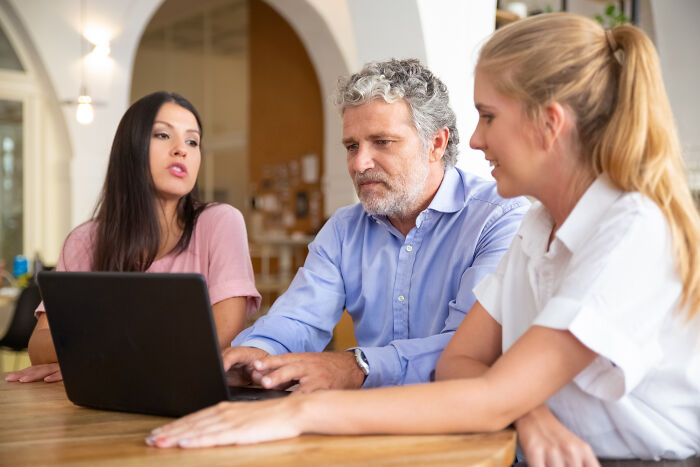 Older man with gray hair discussing aging realities with two young women at a laptop in a casual setting.