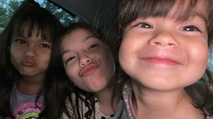 Three smiling children pose closely together in a joyful moment indoors.