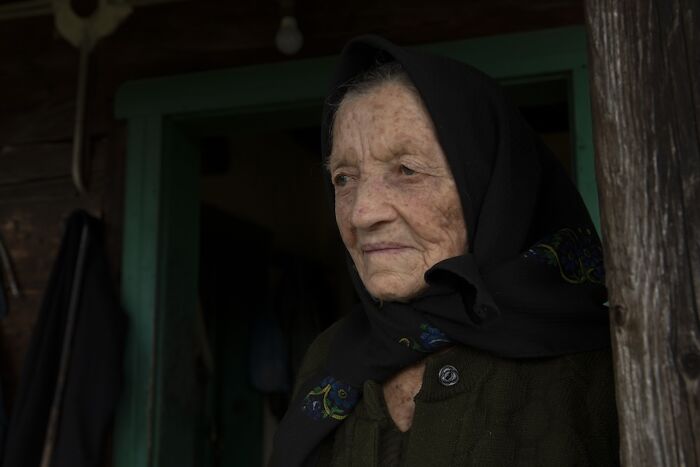 An elderly woman with a headscarf, illustrating vulnerability and aging, stands at a wooden doorway gazing into the distance.