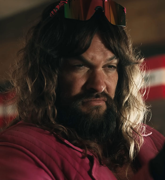 A man with long hair and sunglasses, wearing a pink shirt in a dimly lit room.
