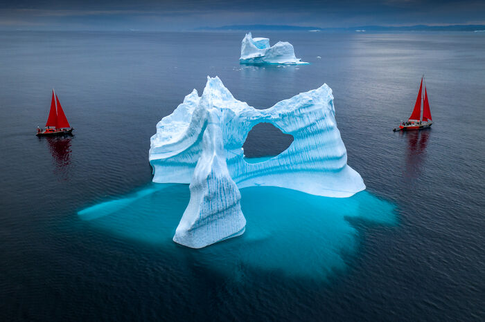 Iceberg with two red-sailed boats in a stunning nature shot.