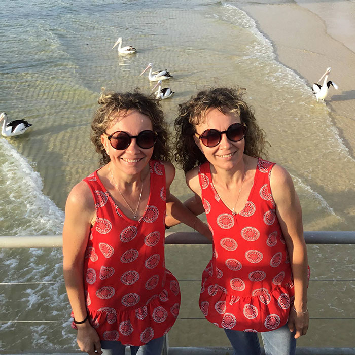 Twins in matching red tops and sunglasses stand by the shore with pelicans nearby. Twins in matching red tops and sunglasses stand by the shore with pelicans nearby.