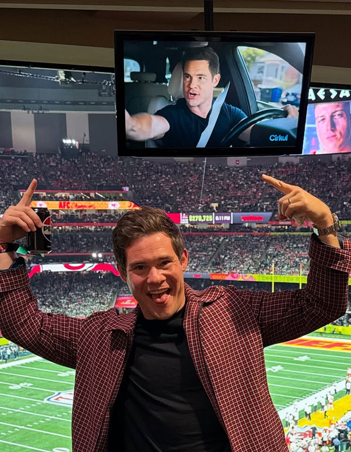 Actor at a football stadium, pointing at a screen showing a scene from a movie.