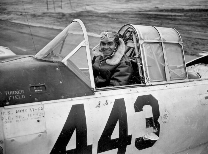 Pilot in WWII cockpit wearing goggles and a flight jacket, sitting in a plane marked with the number 443.