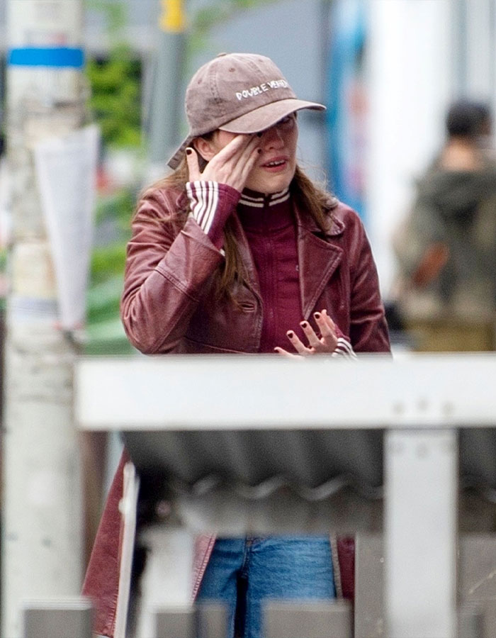 A person in a brown jacket and cap wipes tears while standing on a street, amid recent public drama.