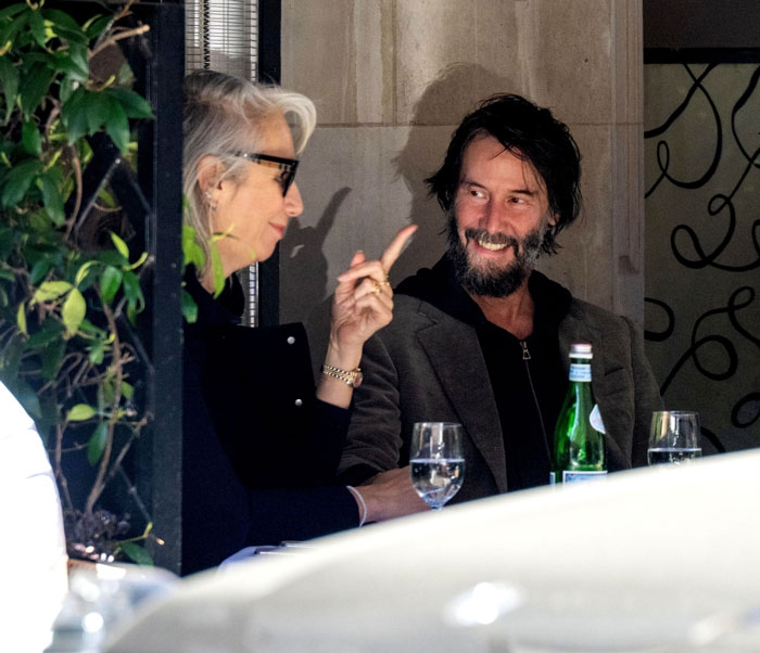 Keanu Reeves with girlfriend at a cafe table, sparking age confusion with his youthful look.
