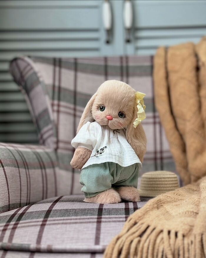 Handmade whimsical plush toy bunny with yellow bow, wearing a white shirt and green pants, sitting on a plaid chair.