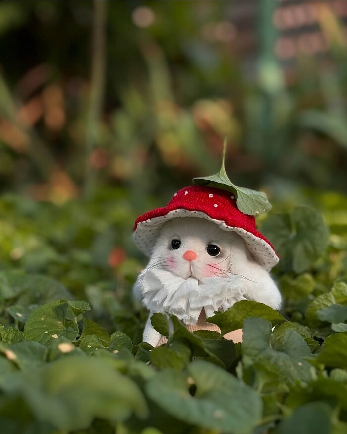 Whimsical plush toy resembling a rabbit, wearing a red mushroom cap, sitting amidst green leaves.