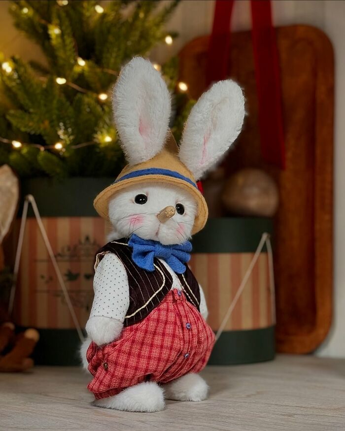 Whimsical plush toy rabbit with a straw hat, red checkered pants, and a blue bow, standing near a decorated tree.