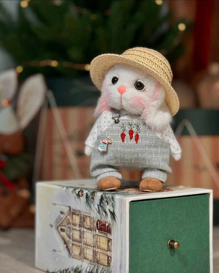Whimsical handmade plush toy rabbit wearing a straw hat and overalls, standing on a decorative box.