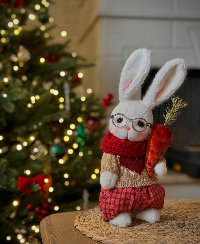 Whimsical handmade plush toy bunny wearing glasses, holding a carrot, with a Christmas tree in the background.