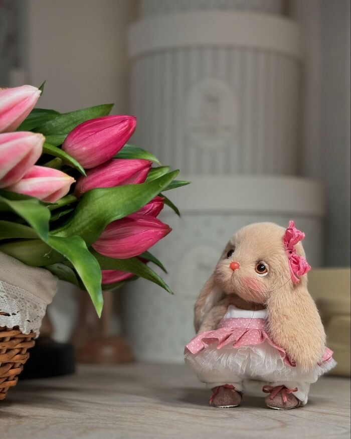 Whimsical plush toy rabbit with pink dress and bow, standing beside a vase of pink tulips.