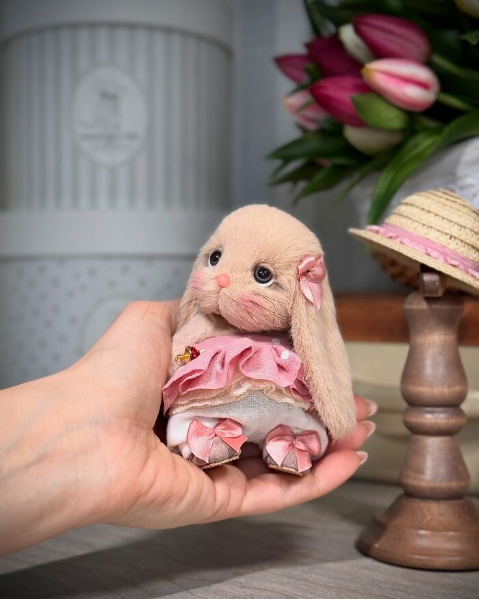 Handmade whimsical plush toy rabbit in a pink dress held in a person's hand, with flowers and a small hat nearby.