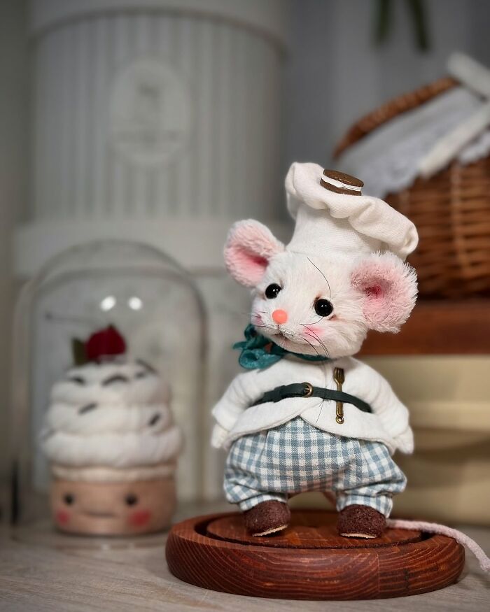 Whimsical handmade plush toy of a mouse chef, wearing a white hat and checkered pants, standing on a wooden base.