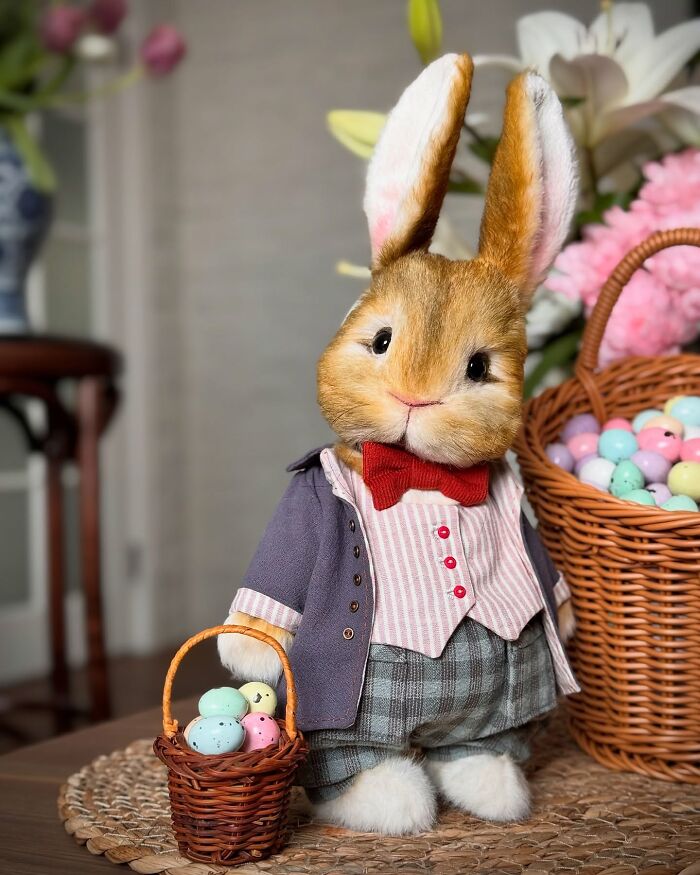 Whimsical plush bunny toy dressed in a suit, holding a small basket of pastel eggs, surrounded by flowers and baskets.