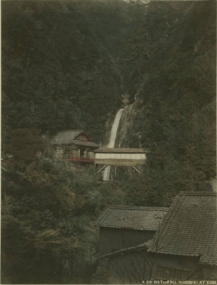 Traditional Japanese buildings near a waterfall surrounded by dense forest, showing Japan in the 1800s.