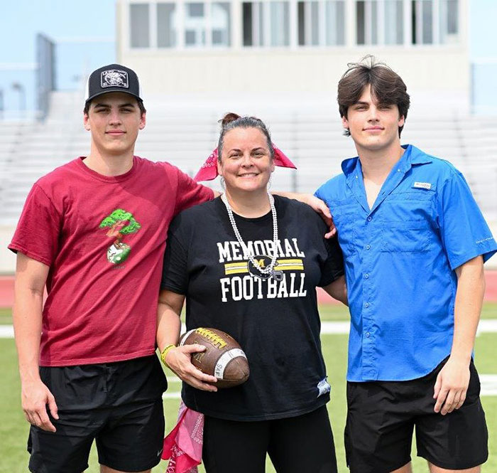 Two brothers and a woman stand on a sports field, one wearing a "Memorial Football" shirt.