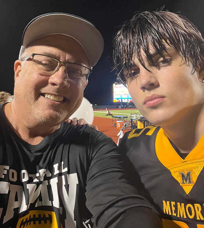 Man in a cap and glasses with a young football player on a field at night.