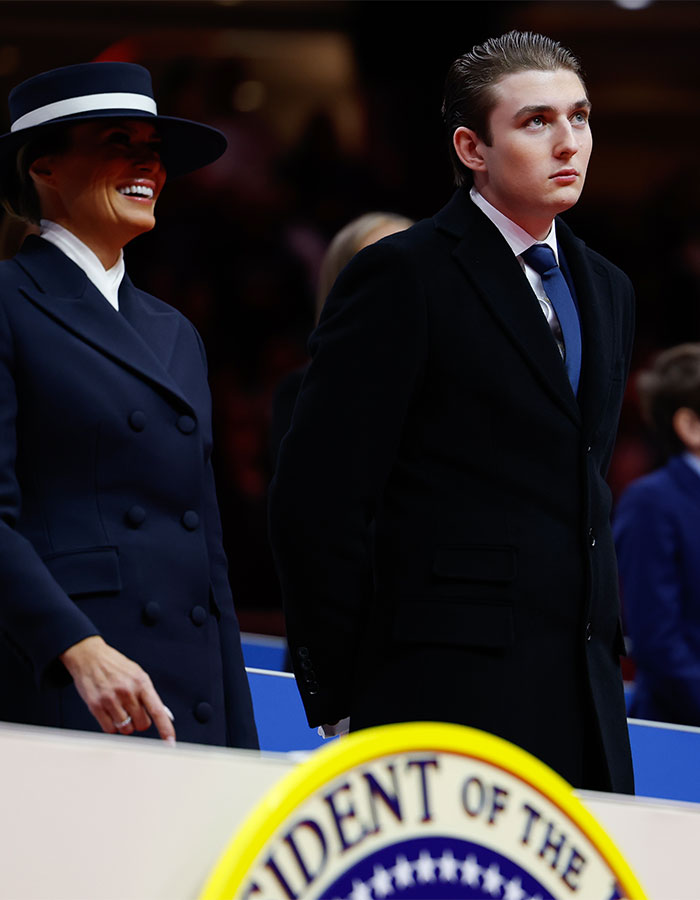 A young man in a dark suit stands beside a smiling woman in a hat, both observing an event with a presidential seal nearby.