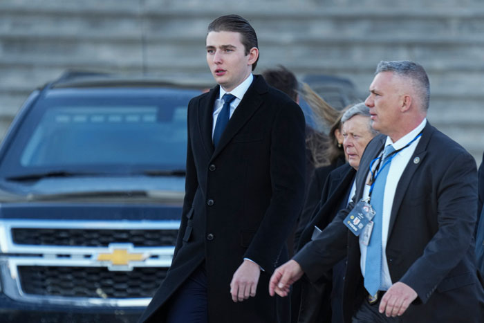 A young man in a coat walking with security personnel on campus.