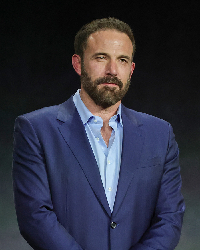 Man in a blue suit standing against a dark background, related to Hollywood scandals.