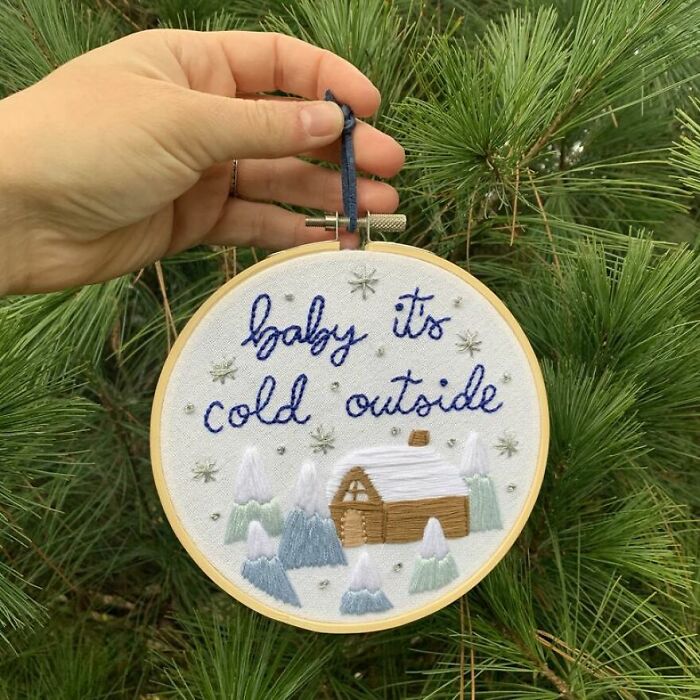 Hand holding a circular embroidery with "baby it's cold outside" text, featuring a winter cabin scene, in front of a pine tree.