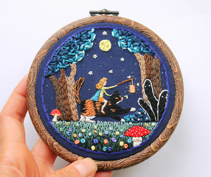 Hand holding hoop with intricate embroidery art showing a whimsical night scene with trees, moon, and a character riding a cat.