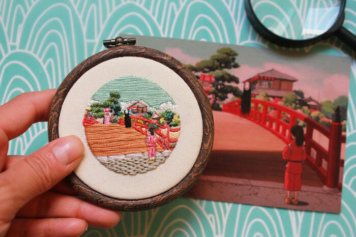 Hand holding an embroidery art piece featuring a traditional Japanese bridge, with a similar scene in the background.