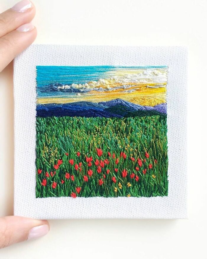 Embroidery art depicting a vibrant landscape with blue skies, mountains, and a field of red flowers.