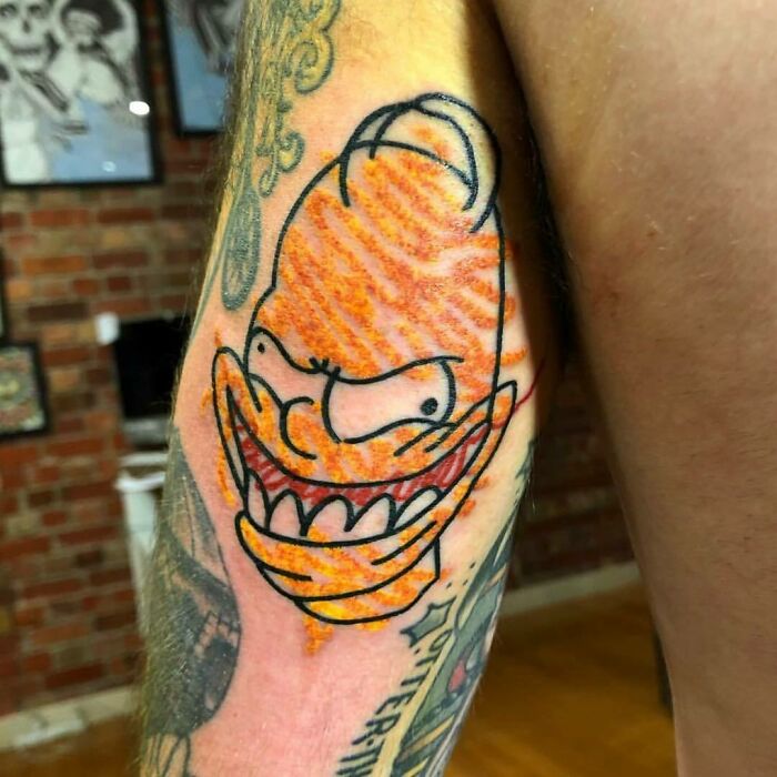 Homer Simpson tattoo on an arm with exaggerated features and vibrant colors, inside a tattoo studio.