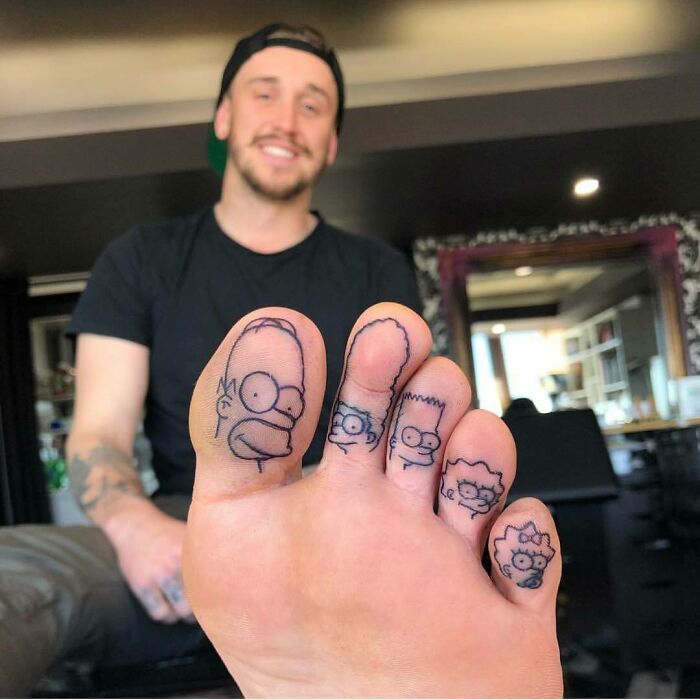 Simpsons tattoos on toes, featuring cartoon characters, with person smiling in the background.