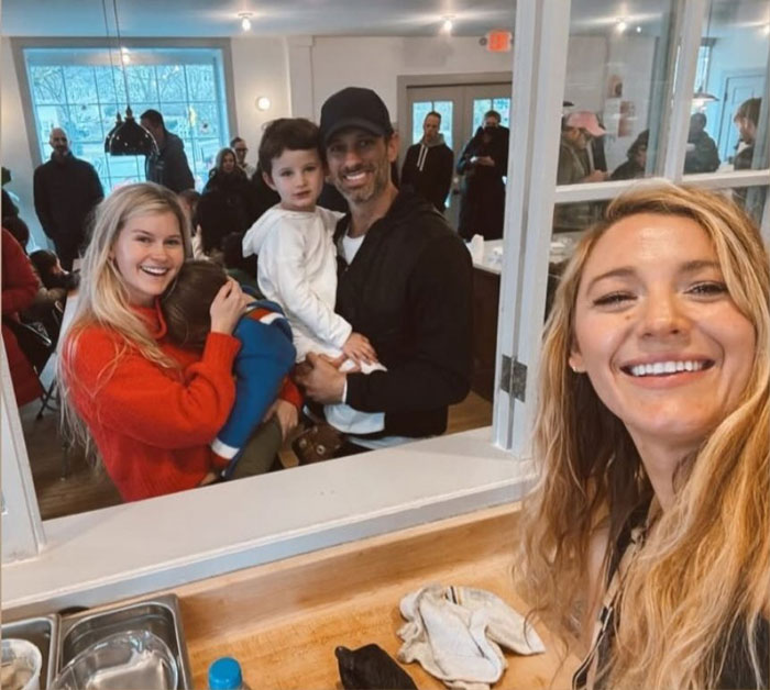 Blonde woman taking a selfie inside a donut shop, smiling with a family in the background.