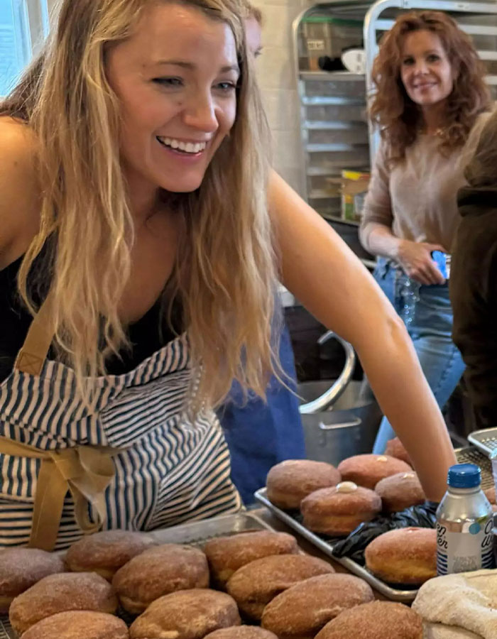 A woman in a striped apron smiling at a donut shop counter with trays of donuts.