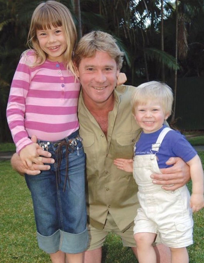 Father with two children outdoors, both smiling and embracing him, in a casual family moment.