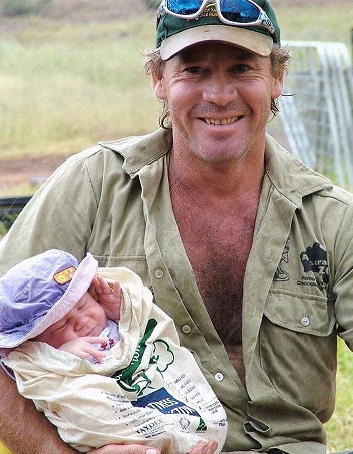 Man smiling in casual attire, holding a newborn swaddled in a cloth.