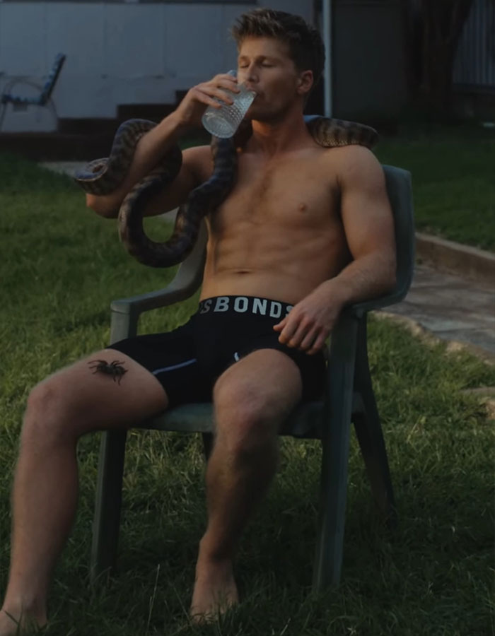 Shirtless man with a snake on his shoulders, sitting outdoors and drinking water.
