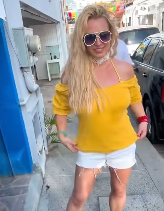 Blonde woman in sunglasses and yellow top, walks confidently on a sunny street, blue and white buildings around her.