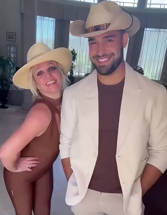Two people in cowboy hats smiling indoors, one in a brown outfit and the other in a beige suit.