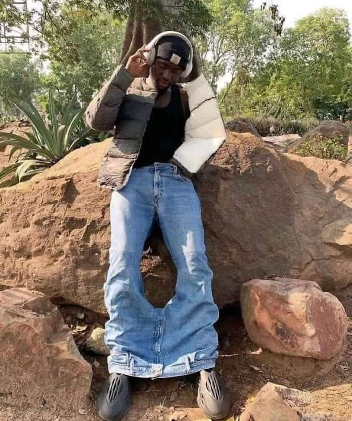 Person in oversized jeans and jacket, standing between rocks, creating a random cursed image in a natural setting.