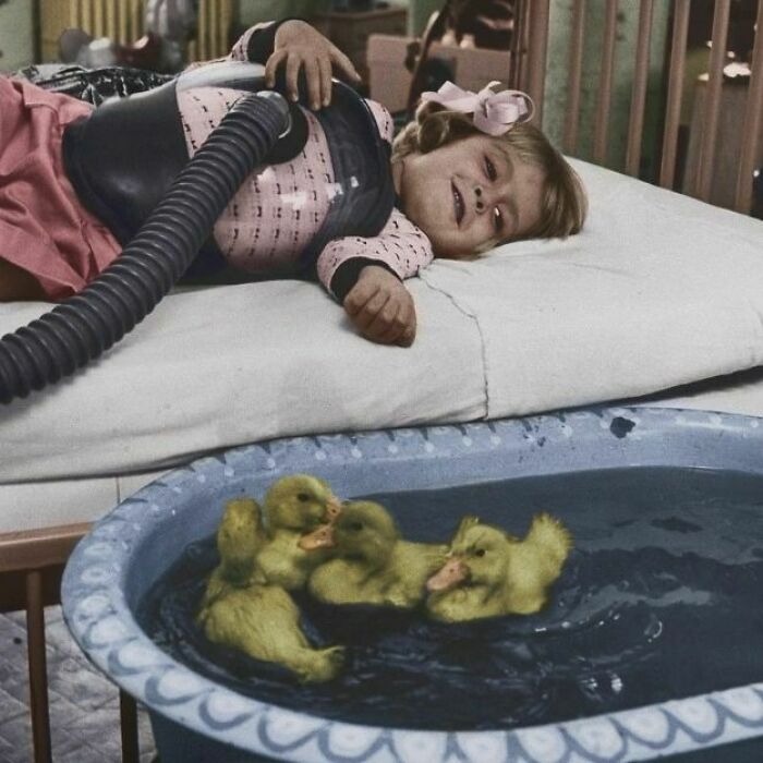 Vintage photo of a child lying in bed with a breathing device, watching ducklings swim in a small tub nearby.