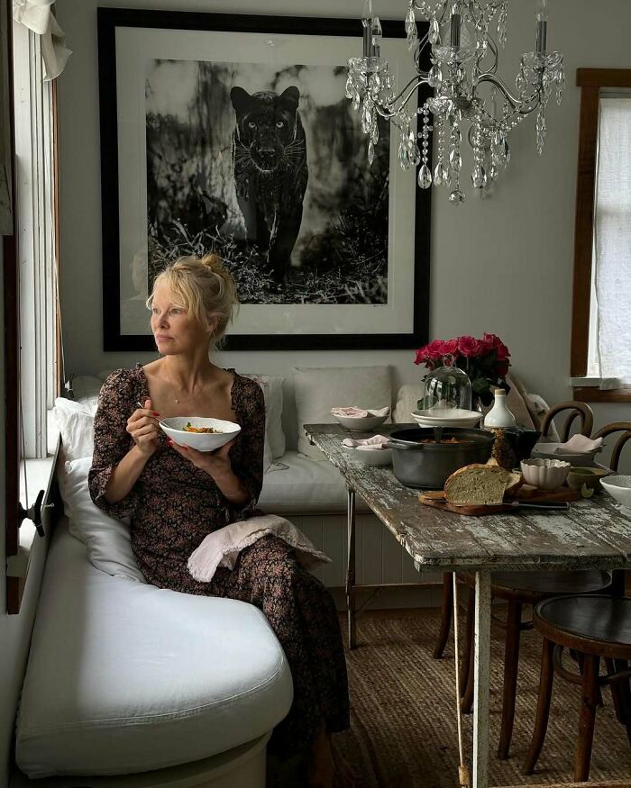 Woman in a floral dress sitting by a window, holding a bowl, in a cozy dining room with rustic decor and celebrity encounters vibe.